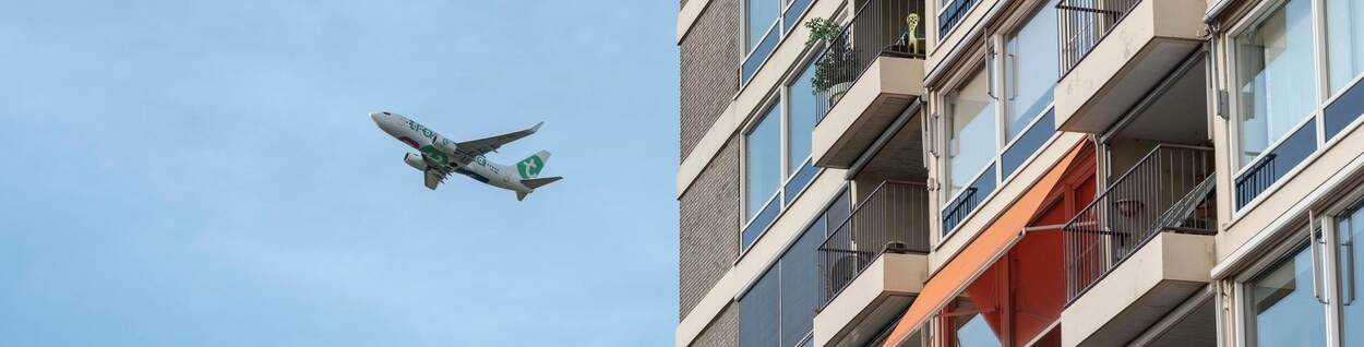 Een vliegtuig vliegt door de lucht. Op de voorgrond staat een flatgebouw.