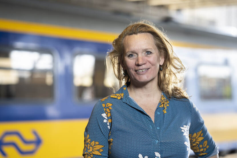 Portretfoto van Margriet van der Goot, haar functie is teamleider Rail. Op de achtergrond staat een blauw gele NS-trein.