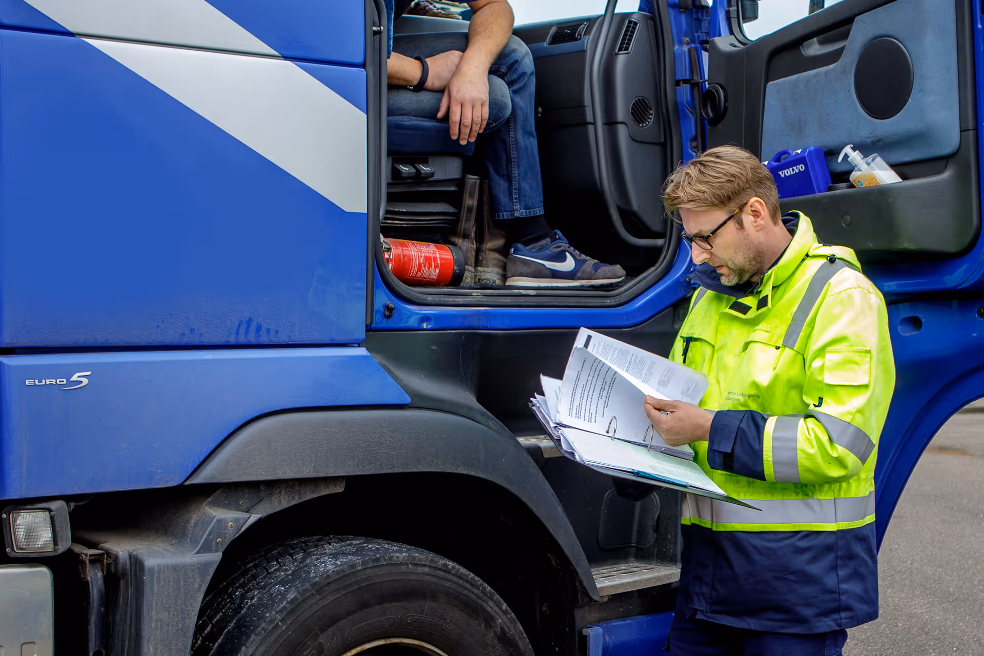 Een inspecteur staat naast een vrachtwagencabine. De chauffeur zit in de cabine. De inspecteur bekijkt enkele papieren.
